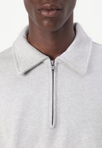 Sweatshirt cinzento com fecho e colarinho, apresentando uma textura suave e um fecho de contraste. O colarinho é ligeiramente estruturado, acrescentando detalhe ao design.
