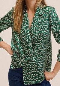 Femme portant un chemisier vert à motif audacieux de maillons de chaîne blancs et noirs, associé à un jean bleu foncé, les mains sur les hanches.