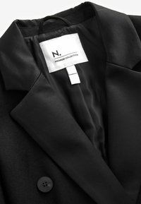 Blazer de la collection premium noire avec col à revers cranté, bouton visible et étiquette intérieure indiquant "N. PREMIUM COLLECTION" sur un tag blanc.