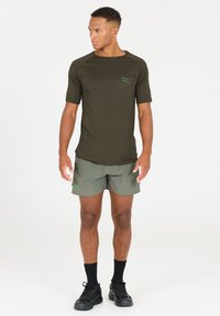 Olivenfarbene Sportshirt mit kurzen Ärmeln, dezenter Musterung und kleinem grünen Logo, kombiniert mit hellgrünen Shorts und schwarzen Laufschuhen.