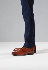 Tan leather dress shoes with a rounded toe, minimal stitching, and a small heel, complemented by navy trousers against a gray background.