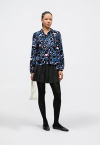 Blå bluse med lange ermer med abstrakte svarte og rosa mønstre, kombinert med et svart plissert skjørt og svarte strømpebukser, holder en hvit veske.