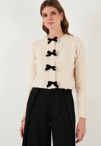 Cardigan fluffy beige chiaro con colletto tondo e cinque fiocchi in velluto nero come bottoni, abbinato a pantaloni neri a vita alta.