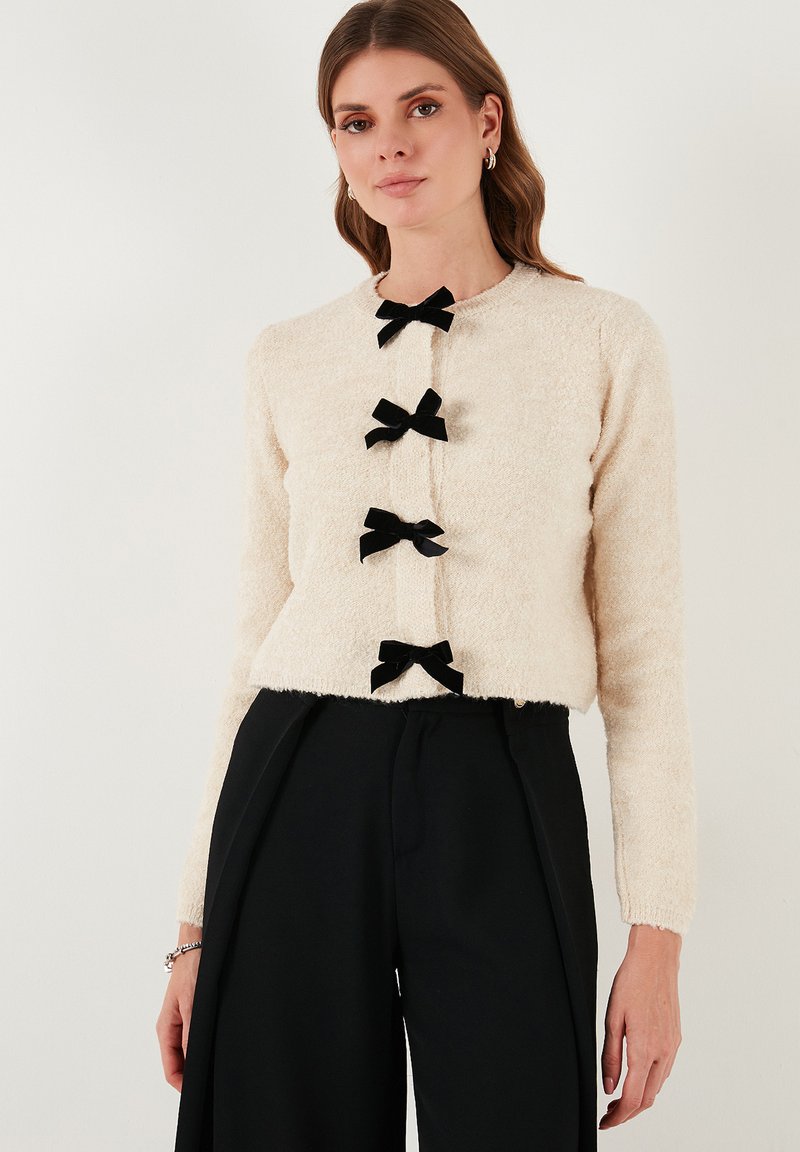 Cardigan fluffy beige chiaro con colletto tondo e cinque fiocchi in velluto nero come bottoni, abbinato a pantaloni neri a vita alta.