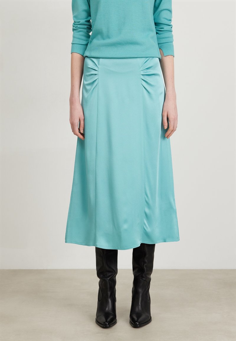 Femme portant une jupe midi turquoise avec des détails froncés aux hanches, associée à un haut à manches longues assorti et des bottes noires montant jusqu'aux genoux.