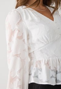 White floral-patterned blouse with sheer, long sleeves; peplum waist; textured fabric; soft, lightweight material with subtle sheen.