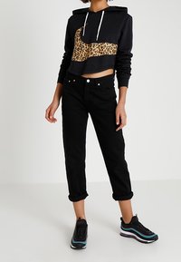 Svart croppad hoodie med leopardmönstrad panel och vita dragsnören; kombinerad med svart högmidjad jeans och svarta sneakers med färgglada detaljer.