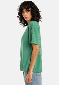 Camiseta de algodón verde con un corte relajado, mangas cortas y un gráfico de texto vertical en el costado. La tela tiene una textura suave.