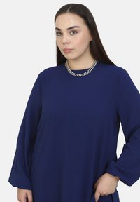 Blaues Langarmkleid mit lockerer Passform, Rundhalsausschnitt und strukturiertem Stoff. Es verfügt über eine silberne Kettenhalskette als Akzentdetail.