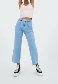 Nainen pukeutuneena vaaleansinisiin korkeavyötäröisiin leveälahkeisiin farkkuihin, vaaleanpunaiseen lyhythihaiseen toppiin ja musta-valkoisiin korkeakorkoisiin tennareihin, seisoo kädet nostettuina.