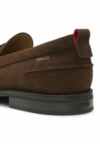 Καφέ καστόρινο παπούτσι slip-on με μαύρη σόλα, κόκκινη θηλιά έλξης και χρυσό λογότυπο "HUGO" στο πλάι κοντά στη φτέρνα.