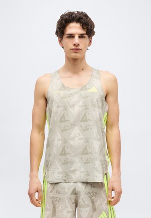 Νεαρός άνδρας φοράει μπεζ αμάνικο αθλητικό τοπ με σχέδιο και αντίστοιχα σορτς με νέον κίτρινες ρίγες Adidas, στέκεται μπροστά σε λευκό φόντο.