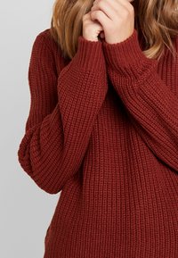 Pull en tricot couleur rouille avec une texture côtelée et des manches longues, doté de poignets ajustés et d'un col large.