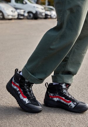 Baskets montantes en camouflage noir et gris, avec une bande rouge, un tissu texturé, une semelle noire robuste avec lacets et languette de tirage.