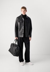 Joven vestido con chaqueta de cuero negra, pantalones negros, zapatillas blancas, sosteniendo un gran bolso de cuero negro, de pie contra un fondo claro y liso.