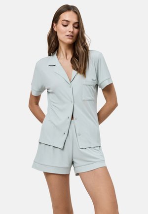 Mujer con camisa de manga corta azul claro abotonada con bolsillo y pantalones cortos a juego, parada con las manos detrás de la espalda sobre fondo blanco.