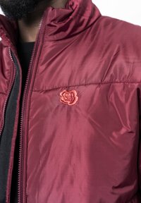 Giubbotto imbottito bordeaux con colletto alto, caratterizzato da un ricamo di una rosa rossa sul petto e una chiusura frontale con zip. Texture liscia.