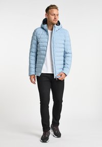 Lyseblå dunjakke med lynlås foran, vandret quiltning og hætte. Sammen med sorte bukser og sporty sorte sko.