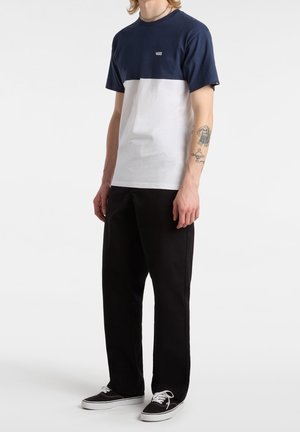 Personne portant un t-shirt Vans bleu marine et blanc, un pantalon noir, des baskets Vans noires, et un tatouage sur l'avant-bras gauche, debout devant un fond blanc.