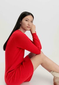 Abito rosso lavorato a maglia con maniche lunghe e silhouette aderente. Texture liscia, lunghezza al ginocchio. Modella seduta con le gambe incrociate, sfondo neutro.
