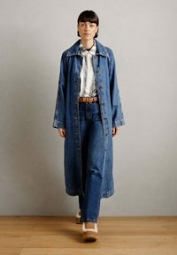 Lange spijkerjas met een kraag, knoopsluiting en zijzakken, gecombineerd met een gestreept blouse en een broek met wijde pijpen. Bruine riem en schoenen.