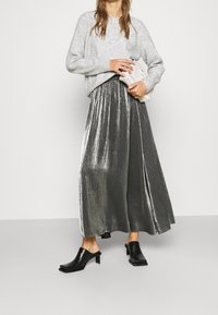 Grå stickad tröja, metallic silver plisserad maxiskärpning och svarta blockklackade mule-skor, hållande en liten vit väska med en tvinnad rem.