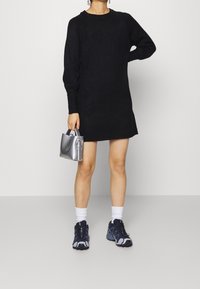 Svart stickad klänning med långa ärmar, texturerade manschetter, parat med en silverfärgad väska och marinblå sneakers, som visar en avslappnad men modern stil.