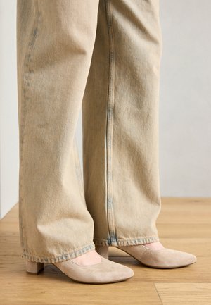 Beige Wildleder-Schuhe mit Blockabsatz und spitzem Schuh, kombiniert mit lockeren, hellbraunen Jeanshosen, die den Fokus auf Textur und Farbkontrast legen.