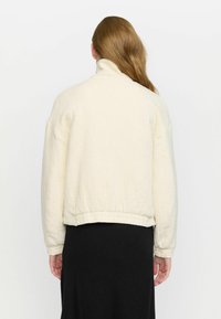 Cream-colored cropped jacket with a textured finish, featuring a high collar and elasticated hem. Worn over a black skirt.