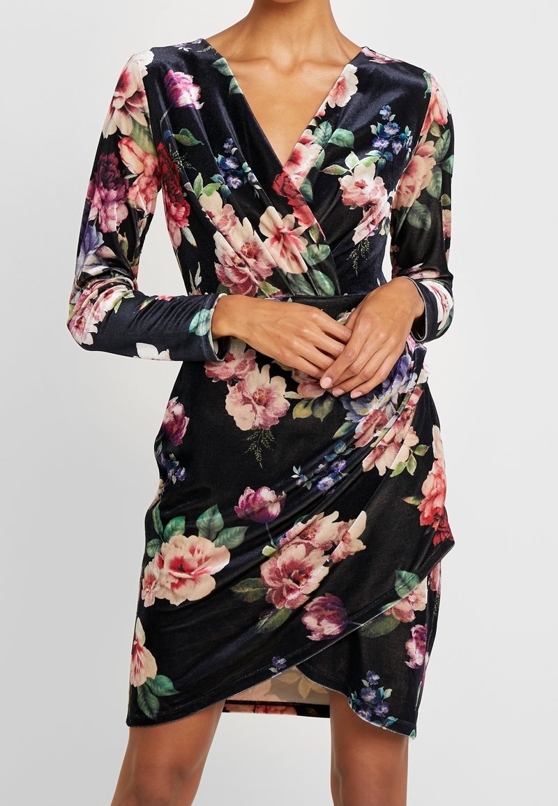 Femme portant une robe portefeuille en velours noir avec un motif floral rose, rouge et violet et des manches longues, les mains jointes à la taille.