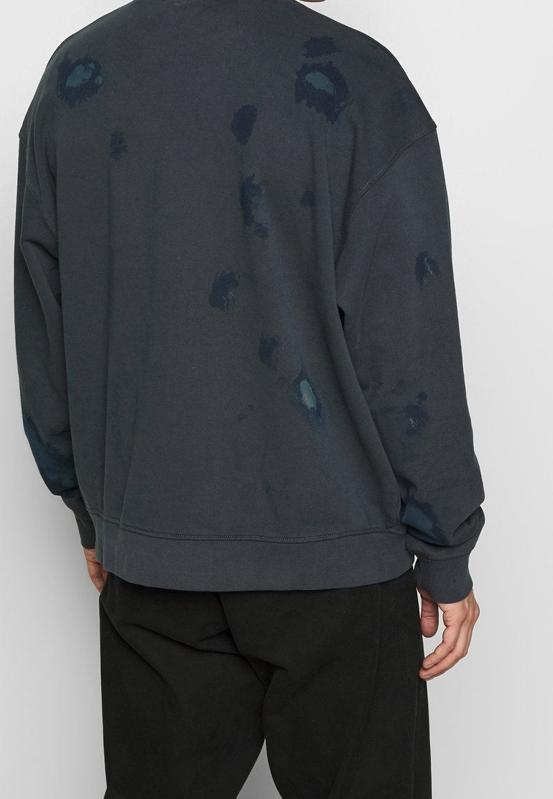 Homme portant un sweat-shirt gris foncé avec des taches décolorées et un pantalon noir, vu de dos sur un fond clair uni.