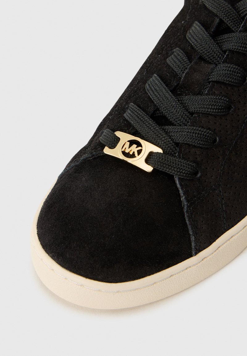 Close-up van een zwart suede sneaker met gouden oogjes en veters, waarbij een textuur bovenwerk en een crèmewitte rubberen zool zichtbaar zijn.