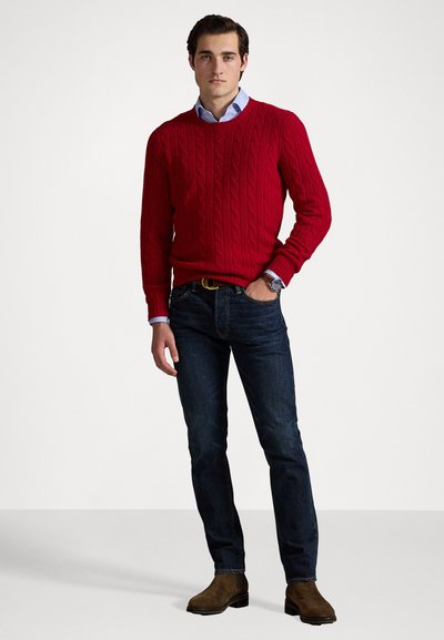 Suéter de punto trenzado rojo, camisa de collar azul claro debajo, jeans azul oscuro, botas de ante marrón, de pie sobre un fondo sencillo.