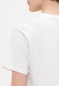 White cotton t-shirt with short sleeves, featuring a crew neckline and smooth texture. The back is partially visible, showing stitching details.