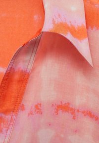 Leichtes, Batikstoff in Orange- und Rosatönen mit glatter Textur. Verfügt über einen Kragen und sichtbare Nähdetails.