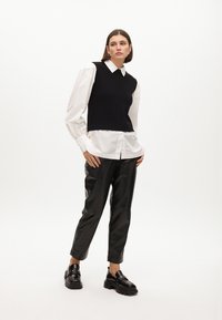 Gilet nero in maglia sovrapposto a una camicia bianca con colletto, abbinato a pantaloni neri lucidi e aderenti e mocassini neri spessi con dettagli dorati.