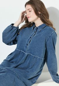 Denimmekko, jossa on kauluksellinen kaula-aukko, nappisulku ja pitkät, puffat hihat. Ominaisuuksiin kuuluu kerroksellinen, rypytetty helma ja pehmeä sininen kankaan rakenne.