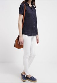 Námornícky krátký rukáv, bílé přiléhavé kalhoty, modré espadrilky s vzorem a hnědá kabelka přes rameno, prezentované v stojící pozici.