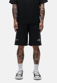 Zwarte katoenen shorts hebben een losse pasvorm met een rechte zoom, logo-details op de linker dij en contrasterende stiksels. Gecombineerd met zwarte sneakers en witte sokken.