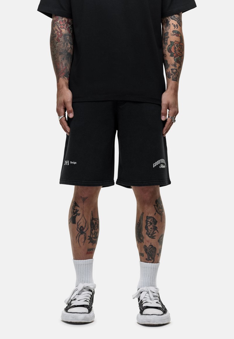 Zwarte katoenen shorts hebben een losse pasvorm met een rechte zoom, logo-details op de linker dij en contrasterende stiksels. Gecombineerd met zwarte sneakers en witte sokken.