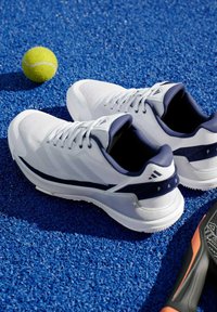 Bílé sportovní tenisky s námořnickými akcenty, síťovinovým svrškem, polstrovanou podrážkou a texturovanou gumovou podešví, umístěné na modrém kurtovém povrchu.