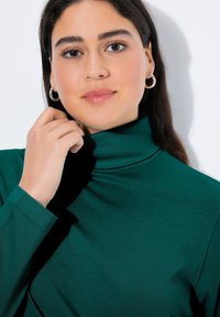 Tmavě zelený rolák s dlouhými rukávy, vyrobený z měkké texturované látky. Má vysoký límec a přiléhavý střih. Viditelné stříbrné kruhové náušnice.