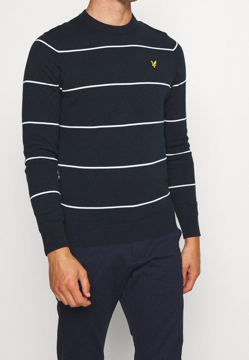 Homme portant un pull bleu marine avec de fines rayures blanches horizontales et un logo oiseau jaune sur la poitrine gauche, associé à un pantalon bleu marine foncé.