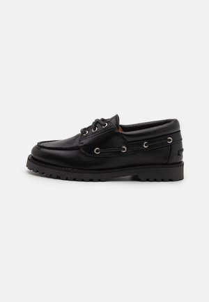 Chaussure bateau en cuir noir avec une semelle épaisse en caoutchouc, un laçage à l'avant, et des œillets métalliques sur un fond clair uni.