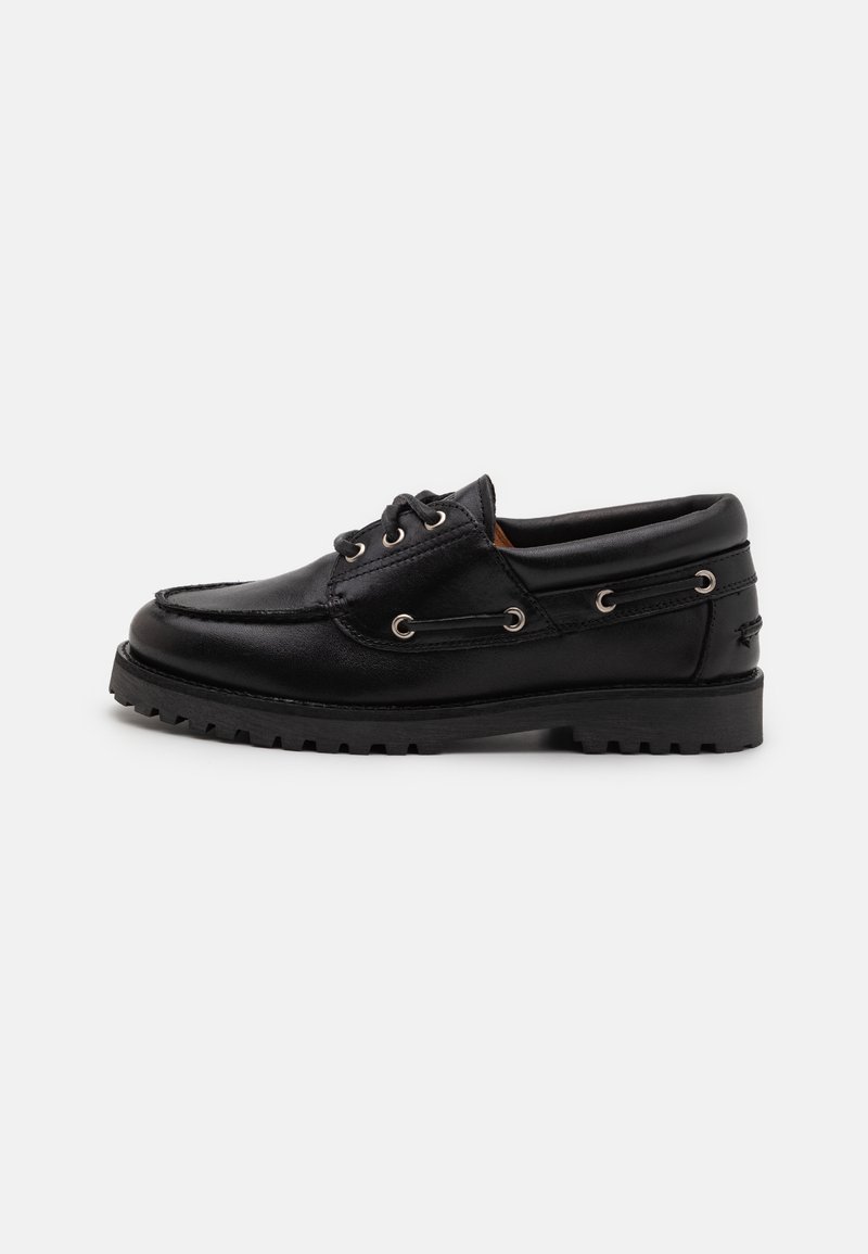 Chaussure bateau en cuir noir avec une semelle épaisse en caoutchouc, un laçage à l'avant, et des œillets métalliques sur un fond clair uni.
