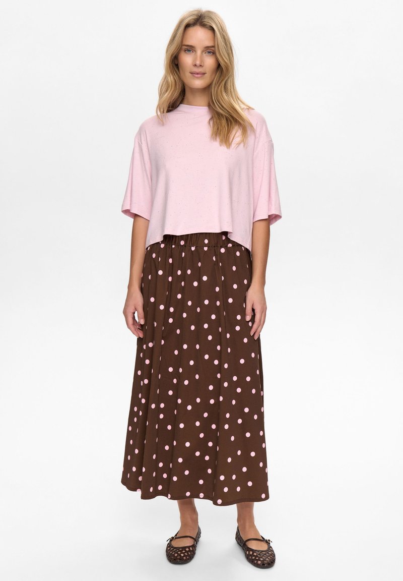 Haut court rose clair à texture lisse, associé à une longue jupe marron à pois roses. Des chaussures noires sans lacets complètent la tenue.