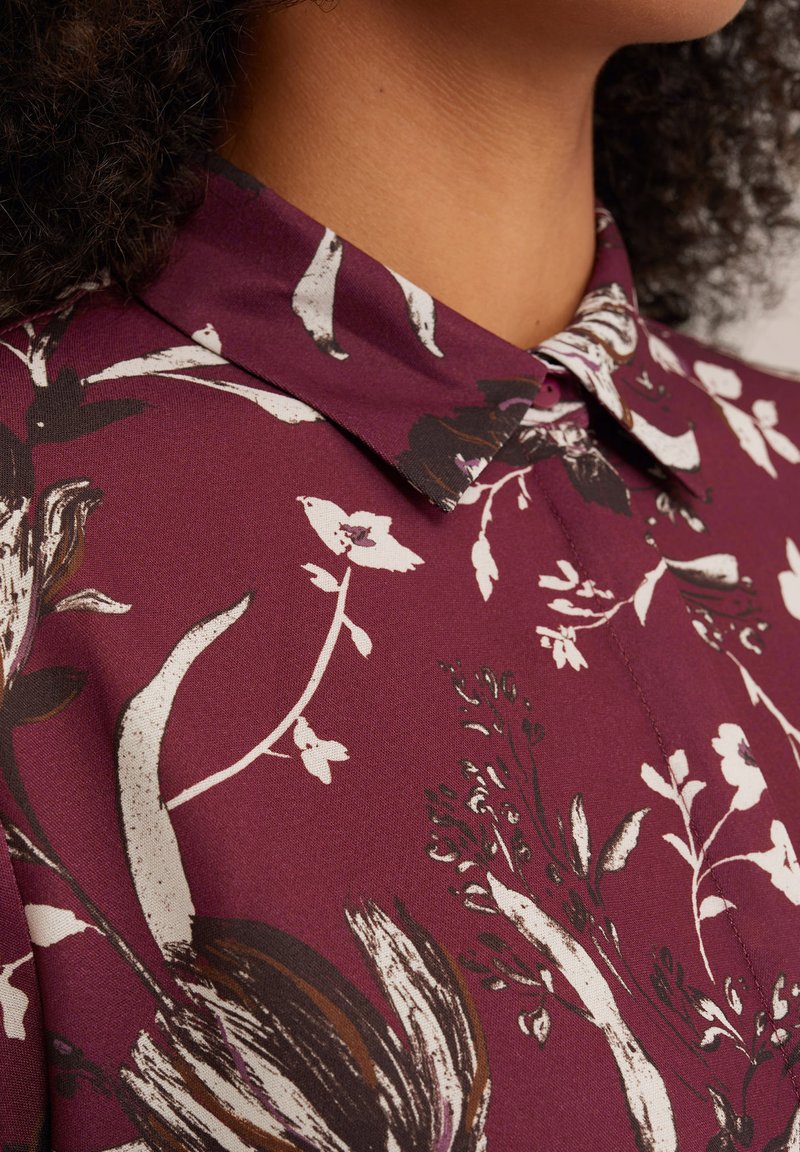 Bluzka z kołnierzykiem w burgundowym materiale z białymi i czarnymi wzorami kwiatowymi. Gładka faktura, z zapięciem na guziki i krótkimi rękawami.