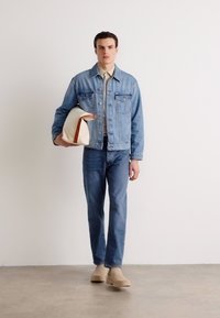 Denim jacket in light blue with pockets, worn over a beige shirt, paired with dark blue jeans and beige boots; holding a cream bag with brown accents.