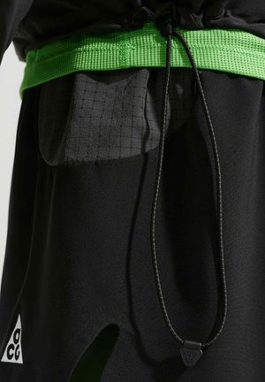 Približna slika črnih hlač z neonsko zelenim pasom, žepom iz mrežastega materiala ter črnim vezalko s trikotno zaponko.