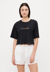 Svart kortärmad t-shirt med färgglad "Calvin Klein" logotyp. Mjuk bomullsmaterial, avslappnad passform med korta ärmar, bärs tillsammans med vita shorts.